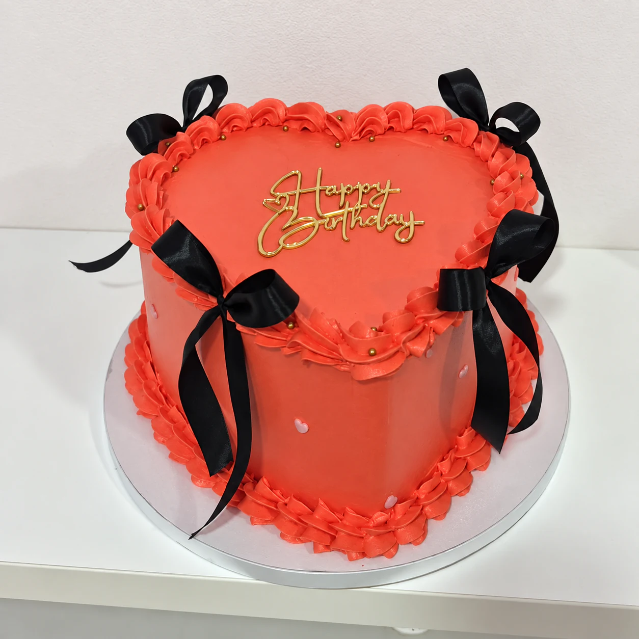 Tarta roja en forma de corazón con bordes de crema, lazos negros y mensaje dorado