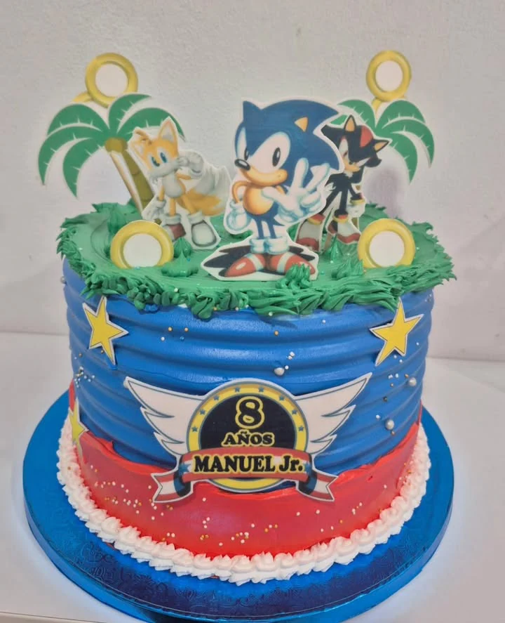 Tarta infantil azul y roja con personajes, anillos, palmeras y césped de crema